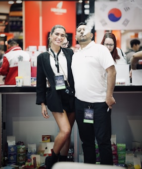 A man and a woman are posing together at an event, both wearing exhibitor badges. The man is exhaling vapor from an e-cigarette, creating a cloud of smoke. The background features signs and a flag, suggesting a diverse or international setting.