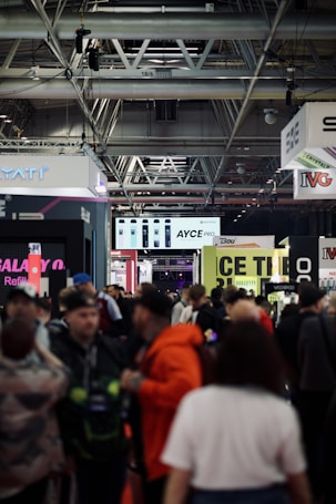 A busy trade show or exhibition with numerous people walking around. Stalls and banners for various brands are visible, with high ceilings showcasing industrial fixtures. Signs above the stalls display brand names and products, creating a bustling and lively atmosphere.