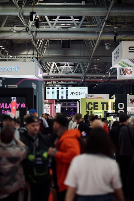A busy trade show or exhibition with numerous people walking around. Stalls and banners for various brands are visible, with high ceilings showcasing industrial fixtures. Signs above the stalls display brand names and products, creating a bustling and lively atmosphere.