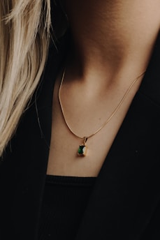 A close-up of a person's neck wearing a gold chain necklace with a small, rectangular green pendant. The person has long, light-colored hair, and is wearing a black top or jacket. The background is a soft focus, enhancing the details of the necklace.