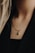 A close-up of a person's neck wearing a gold chain necklace with a small, rectangular green pendant. The person has long, light-colored hair, and is wearing a black top or jacket. The background is a soft focus, enhancing the details of the necklace.