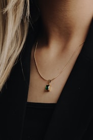 A close-up of a person's neck wearing a gold chain necklace with a small, rectangular green pendant. The person has long, light-colored hair, and is wearing a black top or jacket. The background is a soft focus, enhancing the details of the necklace.