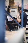 A person stands in a storage space full of shelves holding boxes. They are using a laptop while vaping, with smoke visible. The shelves are filled with various goods, and there's a focus on an 'Ultimate Bar XL' label.