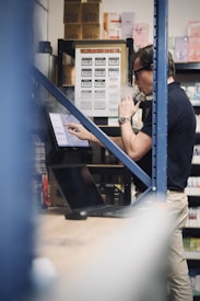 A person stands in a storage space full of shelves holding boxes. They are using a laptop while vaping, with smoke visible. The shelves are filled with various goods, and there's a focus on an 'Ultimate Bar XL' label.