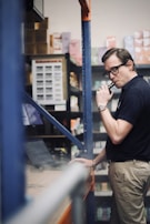 A person in a warehouse setting is looking at a laptop while holding a vape device. Shelves are stocked with various boxes and products in the background.
