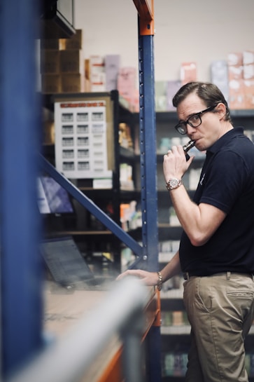 A person in a warehouse setting is looking at a laptop while holding a vape device. Shelves are stocked with various boxes and products in the background.