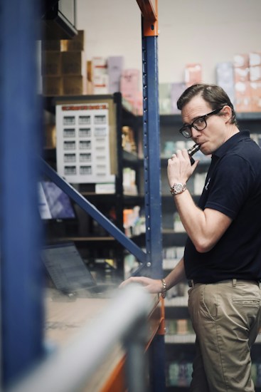A person in a warehouse setting is looking at a laptop while holding a vape device. Shelves are stocked with various boxes and products in the background.