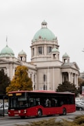 A large, red public bus is driving along a city street with a historical building featuring green domes in the background. The sky is overcast, and there are some autumn trees with yellow leaves nearby.