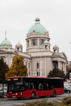A large, red public bus is driving along a city street with a historical building featuring green domes in the background. The sky is overcast, and there are some autumn trees with yellow leaves nearby.