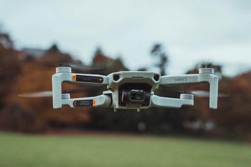 A compact drone with a sleek design hovers in mid-air, displaying its propellers and camera prominently. The background features a blurred natural setting, suggesting it is flying outdoors.