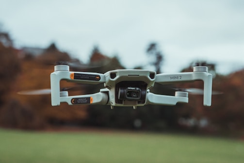 A compact drone with a sleek design hovers in mid-air, displaying its propellers and camera prominently. The background features a blurred natural setting, suggesting it is flying outdoors.