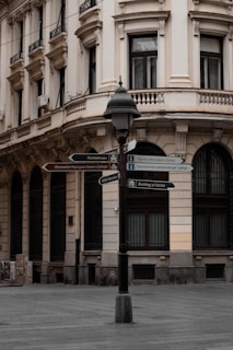 An urban street scene in front of a classic, multi-story building with ornate architectural details. The building has large windows with black grills, balconies, and cream-colored walls. A lamppost stands prominently in the foreground, with multiple signs pointing in various directions, indicating tourist attractions and locations in both English and another language (likely Serbian). The street appears empty and there is minimal activity.