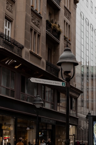 A street scene featuring an old building with ornate architectural details and balconies, contrasted by a modern glass building in the background. A street lamp and a sign for the Tourism Organisation of Serbia are prominently visible, with several people walking nearby.