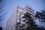 A modern, multi-story building with a distinctive curved architecture, adorned with a large vertical banner promoting an event called 'Seoul Design'. The building is surrounded by tall trees, and the sky is a clear dusk blue, giving the scene an early evening ambiance.