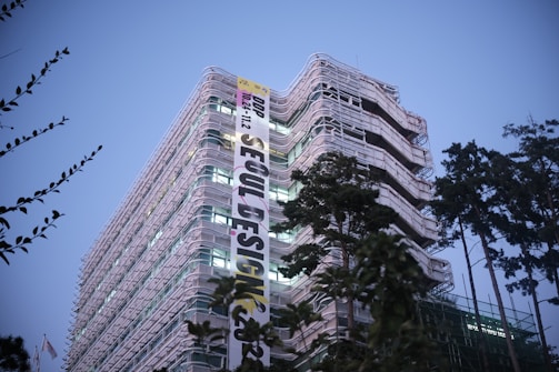 A modern, multi-story building with a distinctive curved architecture, adorned with a large vertical banner promoting an event called 'Seoul Design'. The building is surrounded by tall trees, and the sky is a clear dusk blue, giving the scene an early evening ambiance.