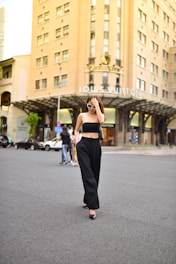 A person wearing sunglasses and a stylish black outfit walks confidently in an urban setting. The background features a prominent building with 'Louis Vuitton' signage, indicating a high-end retail area. Other pedestrians and parked cars are also visible.