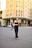 A person wearing sunglasses and a stylish black outfit walks confidently in an urban setting. The background features a prominent building with 'Louis Vuitton' signage, indicating a high-end retail area. Other pedestrians and parked cars are also visible.