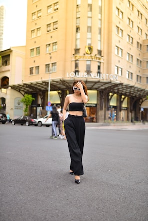 A person wearing sunglasses and a stylish black outfit walks confidently in an urban setting. The background features a prominent building with 'Louis Vuitton' signage, indicating a high-end retail area. Other pedestrians and parked cars are also visible.
