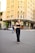 A person wearing sunglasses and a stylish black outfit walks confidently in an urban setting. The background features a prominent building with 'Louis Vuitton' signage, indicating a high-end retail area. Other pedestrians and parked cars are also visible.