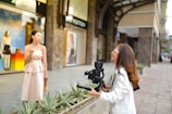 Outside a stylish storefront, a woman in a light-colored dress stands in front of a camera held by another woman wearing a white jacket. They appear to be engaged in filming or photography. The background includes a display window with mannequins and colorful decor, suggesting a city shopping area.