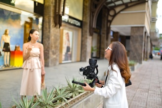 Outside a stylish storefront, a woman in a light-colored dress stands in front of a camera held by another woman wearing a white jacket. They appear to be engaged in filming or photography. The background includes a display window with mannequins and colorful decor, suggesting a city shopping area.