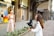 Outside a stylish storefront, a woman in a light-colored dress stands in front of a camera held by another woman wearing a white jacket. They appear to be engaged in filming or photography. The background includes a display window with mannequins and colorful decor, suggesting a city shopping area.