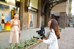 Outside a stylish storefront, a woman in a light-colored dress stands in front of a camera held by another woman wearing a white jacket. They appear to be engaged in filming or photography. The background includes a display window with mannequins and colorful decor, suggesting a city shopping area.