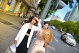 Two women are walking on a city sidewalk. One is wearing a white jacket with a black crop top and wide-leg pants, holding a shopping bag. The other is dressed in a beige dress, holding a wide-brimmed straw hat. They are surrounded by city buildings and parked motorbikes, with greenery and cars visible on the road beside them.
