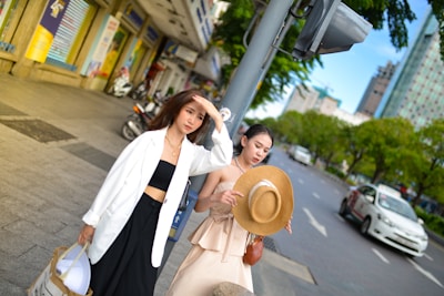 Two women are walking on a city sidewalk. One is wearing a white jacket with a black crop top and wide-leg pants, holding a shopping bag. The other is dressed in a beige dress, holding a wide-brimmed straw hat. They are surrounded by city buildings and parked motorbikes, with greenery and cars visible on the road beside them.
