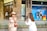 A woman in an elegant beige dress and straw hat stands in front of a Louis Vuitton store, while another person in a white outfit films her with a professional camera. Mannequins are visible in the store window behind them.