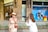A woman in an elegant beige dress and straw hat stands in front of a Louis Vuitton store, while another person in a white outfit films her with a professional camera. Mannequins are visible in the store window behind them.