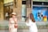 A woman in an elegant beige dress and straw hat stands in front of a Louis Vuitton store, while another person in a white outfit films her with a professional camera. Mannequins are visible in the store window behind them.