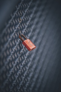 A red padlock with a heart symbol is securely fastened to a chain-link fence, conveying a sense of intimacy or affection amidst a cold, industrial backdrop.
