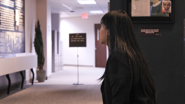 A woman with long dark hair is standing in a hallway or gallery, looking at a display board mounted on the wall. The environment is well-lit, with a potted plant nearby. There are various framed objects and signs visible, and the space has a professional or institutional appearance.