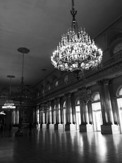 An opulent hall featuring large, ornate chandeliers hanging from a high ceiling. The space is illuminated by natural light streaming through tall windows, casting shadows on the polished floor. The room is surrounded by tall columns and an intricately designed balcony railing.
