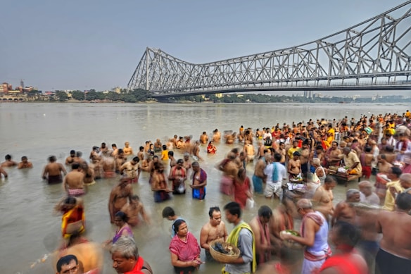 A large group of people gathered in the river near a massive steel bridge, possibly participating in a religious or cultural ritual. Many are partially submerged in the water, some holding offerings, wearing traditional attire. The sky is overcast, and buildings are visible in the distance across the river.