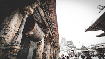 A large, ancient stone structure with intricate carvings is prominently featured. The architecture includes tall columns and detailed sculptures. In the background, a temple tower, or gopuram, rises, showcasing traditional design elements. The sky is overcast, creating a dramatic backdrop.
