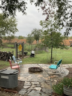 A backyard setting with a round stone patio that features a fire pit in the center. Surrounding the fire pit are four colorful metal chairs with various cushions. The area is surrounded by green grass, trees, and a wooden fence. There's a sign on the fence that reads 'Pavement Ends'. A wheelbarrow is visible near the fence, and a stack of firewood is piled up on the left side.
