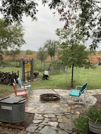 A backyard setting with a round stone patio that features a fire pit in the center. Surrounding the fire pit are four colorful metal chairs with various cushions. The area is surrounded by green grass, trees, and a wooden fence. There's a sign on the fence that reads 'Pavement Ends'. A wheelbarrow is visible near the fence, and a stack of firewood is piled up on the left side.