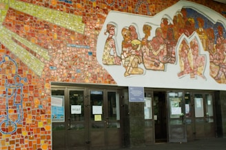 A colorful mural with mosaic tiles depicting abstract human figures sits above a set of glass doors. The mosaic features vibrant reds, yellows, blues, and greens, creating an intricate pattern. The figures appear to be interacting with each other, surrounded by a dynamic background.