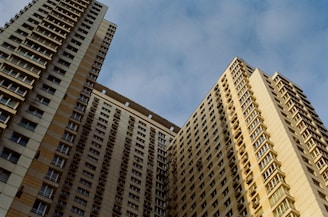 Two high-rise apartment buildings with numerous windows and balconies are positioned closely together, creating a towering perspective against a blue, slightly cloudy sky.