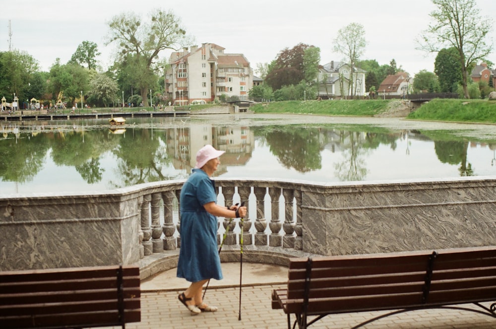 a woman in a blue dress and a white hat is walking by a lake