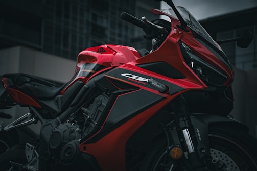 A sleek, red motorcycle is parked outdoors with water droplets visible on its surface, indicating recent rain. The image captures the precise, aerodynamic design and the intricate details of the engine and bodywork.