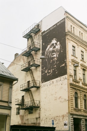 A large mural on a multistory building wall with a striking portrait of a theatrical figure. The mural includes text in a different language. The building has a metal fire escape attached to its side and exhibits an aged, weathered facade.