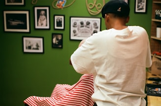 A person wearing a white shirt and black cap is standing in a room decorated with several framed photographs and artwork on the walls. The person appears to be cutting hair or grooming someone seated in front of them, covered with a red and white striped cloth. The room has a green wall and some shelves on the side.