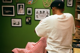 A person wearing a white shirt and black cap is standing in a room decorated with several framed photographs and artwork on the walls. The person appears to be cutting hair or grooming someone seated in front of them, covered with a red and white striped cloth. The room has a green wall and some shelves on the side.