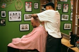 A barber wearing a cap is giving a haircut to a customer draped in a red and white striped cape. The setting is a barbershop with green walls adorned with various framed photographs and memorabilia.