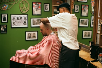 A barber wearing a cap is giving a haircut to a customer draped in a red and white striped cape. The setting is a barbershop with green walls adorned with various framed photographs and memorabilia.