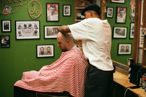 A barber wearing a cap is giving a haircut to a customer draped in a red and white striped cape. The setting is a barbershop with green walls adorned with various framed photographs and memorabilia.