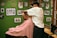 A barber wearing a cap is giving a haircut to a customer draped in a red and white striped cape. The setting is a barbershop with green walls adorned with various framed photographs and memorabilia.